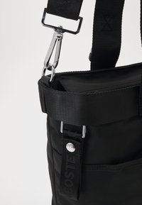 Borsa in tessuto nero con superficie testurizzata, dotata di una clip in metallo argentato, una tracolla e un'etichetta nera con marchio e scritta bianca.