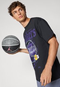 Sort grafisk t-shirt med teksten "Los Angeles Lakers" i lilla, basketballgrafik og et rødt Jordan-logo. Holder en struktureret basketball.