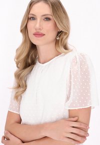 Hvid bluse med teksturerede polka-prikker, gennemsigtige puffærmer og rund halsudskæring. Blødt stof med diskret drapering og en delikat finish.