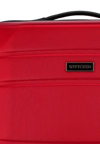 Valigia rigida rossa con finitura testurizzata, dotata di una maniglia nera e di un prominente logo nero del marchio. Linee orizzontali aggiungono dettaglio.