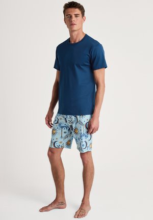 Marineblaues T-Shirt mit Rundhalsausschnitt kombiniert mit hellblauen Badeshorts, die Sonnen- und Mondmuster aufweisen. Das Outfit ist lässig und eignet sich gut als Strandbekleidung.