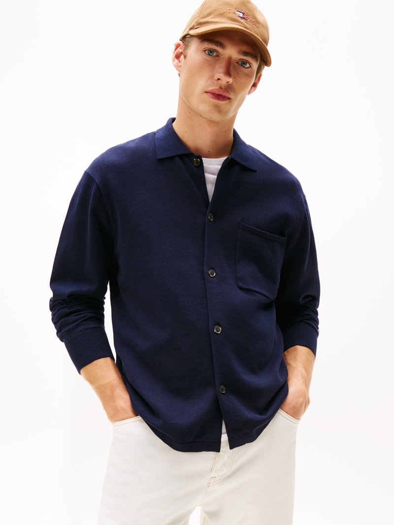 Cardigan lavorato a maglia blu navy con colletto, bottoni frontali e taschino sul petto. Il modello indossa una maglietta bianca e pantaloni beige. Texture liscia.