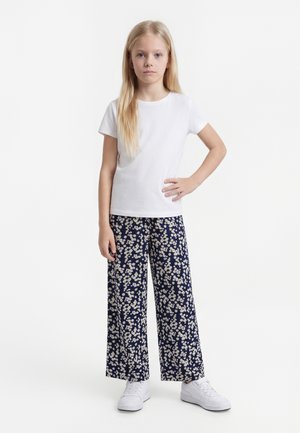 Fille blonde debout, main sur la hanche, portant une chemise blanche à manches courtes, un pantalon large bleu marine à imprimé floral blanc, et des baskets blanches.