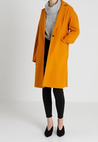 Senfgelber, langer Wollmantel mit Reverskragen, zwei seitlichen Taschen und lässiger Passform, kombiniert mit einem grauen Strickpullover und schwarzen Leggings.