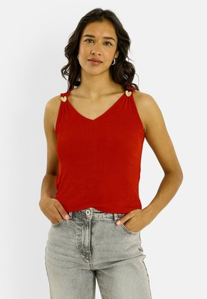 Jeune femme aux cheveux bruns ondulés portant un haut rouge sans manches avec des détails en forme de cœur sur les bretelles et un jean gris clair, les mains dans les poches.