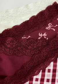 Spetskanter i vinröd och vit, med blommönster och rosetter, lager på olika texturerade tyger, inklusive rutigt och enfärgat.