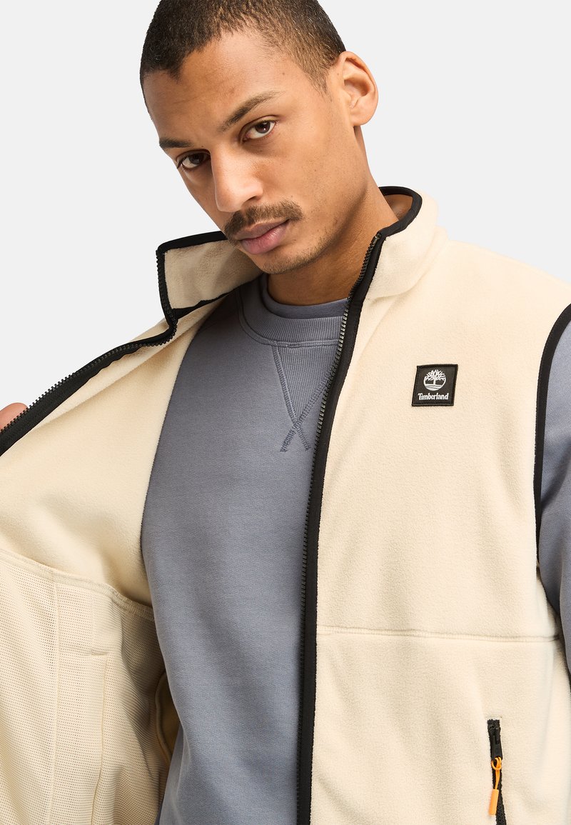 Giacca in pile beige chiaro con accenti e cerniere nere, dotata di una toppa con il logo. Indossata aperta sopra una felpa grigia. Tessuto texture.