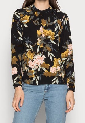 Kvinde iført en sort langærmet top med gule og pink blomsterprint kombineret med lyseblå jeans.