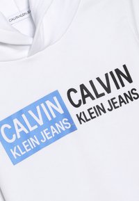 Biała bluza z kapturem z niebieskim prostokątem, na którym znajduje się napis "CALVIN" oraz "KLEIN JEANS" w czerni, wykonana z miękkiego materiału o gładkiej fakturze.