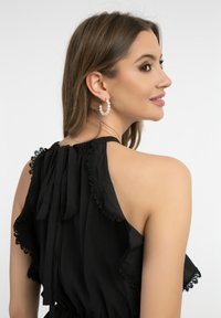 Schwarze Bluse mit einem Rüschendesign und Spitzeneinsätzen, Neckholder-Ausschnitt und einer Schleife auf der Rückseite. Accessoires mit Perlen-Hoops-Hängern.
