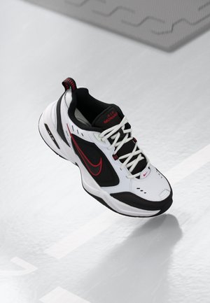 Baskets Nike Air Monarch blanches, noires et rouges avec lacets blancs, logo sur la semelle intermédiaire et Swoosh rouge vues de dessus sur fond de sol clair.
