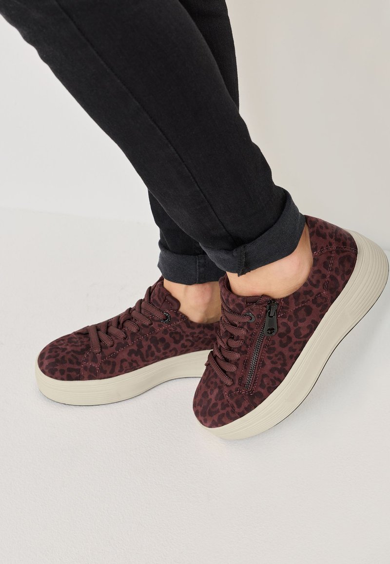 Baskets en daim bordeaux avec un motif léopard, lacets texturés, semelle plateforme et détail de fermeture éclair sur le côté, associées à un jean noir retroussé.