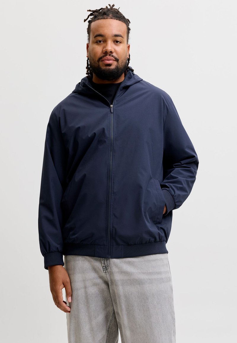 Homme aux cheveux tressés portant une veste zippée bleu marine et un jean gris clair, debout devant un fond clair uni.