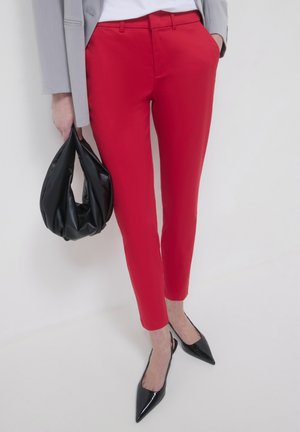 Red tailored pants with slim fit, smooth fabric, mid-rise waist, and belt loops. Paired with black pointed-toe heels and a black bag.