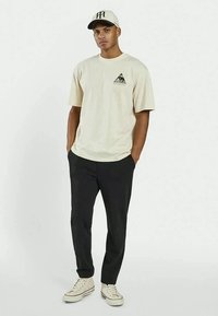 Beige bomulds t-shirt med korte ærmer, med et trekantsgrafik på venstre bryst. Kombination med sorte bukser og lyse sneakers.