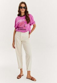 Růžové bavlněné tričko s grafickým potiskem, spárované se smetanově zbarvenými, mírně zúženými kalhotami. Hnědé skládací sandály s designem křížových popruhů.