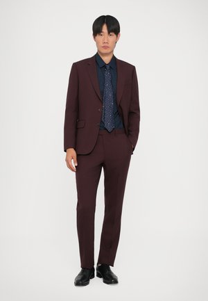 Costume bordeaux à coupe ajustée, avec des revers à cran et deux boutons. Porté avec une chemise sombre et une cravate à motifs. Des chaussures de ville noires complètent le look.