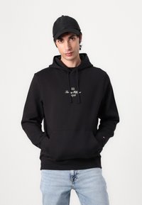 Sudadera negra con un bolsillo frontal, capucha con cordones y texto bordado. Llevada sobre una camiseta negra, combinada con jeans azul claro.