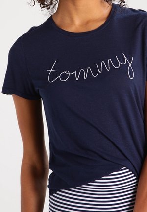 Frau trägt ein marineblaues T-Shirt mit weißer, in Schreibschrift gestickter Aufschrift "tommy" und einen gestreiften Rock in Marineblau und Weiß.