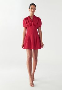 Rotes kurzes Kleid mit puffigen Ärmeln, Knopfkragen und tailliertem Schnitt. Ausgestellter Rock und glattes Material. Ein Paar Riemchensandalen.