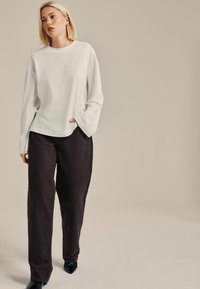 Witte blouse met lange mouwen in een losse pasvorm, gecombineerd met donkere, wijde broek en zwarte puntige schoenen, voor een neutrale achtergrond.