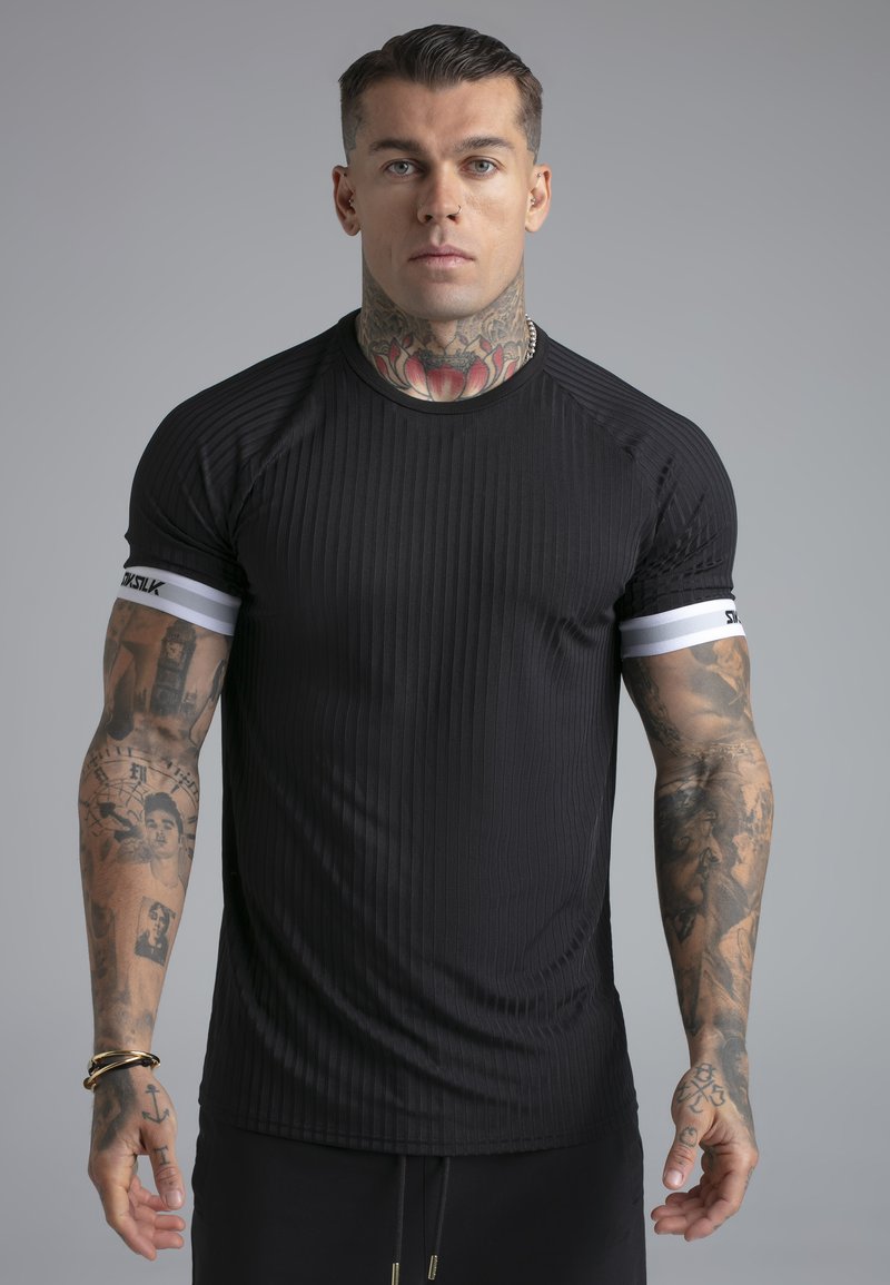 Homme aux bras et au cou tatoués, portant un t-shirt noir côtelé à manches courtes avec des poignets à rayures blanches, debout devant un fond gris.