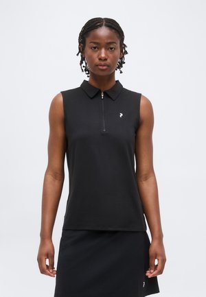 Kvinde med flettet hår iført en ærmeløs sort polo-top med lynlås og et matchende sort nederdel mod en ensartet hvid baggrund.