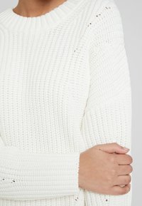 Maglione bianco lavorato a maglia con texture a coste, con scollo rotondo e maniche lunghe. Dettagli in punto aperto lungo le cuciture.