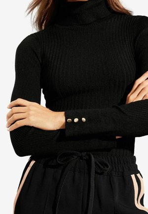 Personne portant un pull à col roulé côtelé noir avec trois boutons métalliques sur le poignet et un pantalon noir avec des bandes latérales claires et un cordon de serrage.