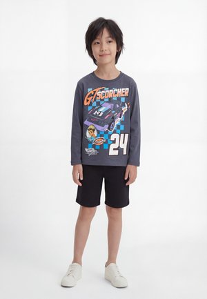 Enfant aux cheveux foncés portant un t-shirt gris à manches longues avec un dessin de voiture de course et un short noir, debout sur un fond blanc.