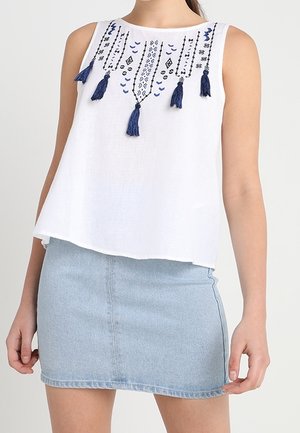 Haut blanc sans manches avec des motifs bleus brodés et des pompons, associé à une mini-jupe en denim bleu clair.