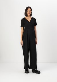 Combinaison noire avec un haut style cache-coeur et des manches courtes, présentant un pantalon large, fabriquée à partir d'un tissu lisse, associée à des chaussures noires chunkys.