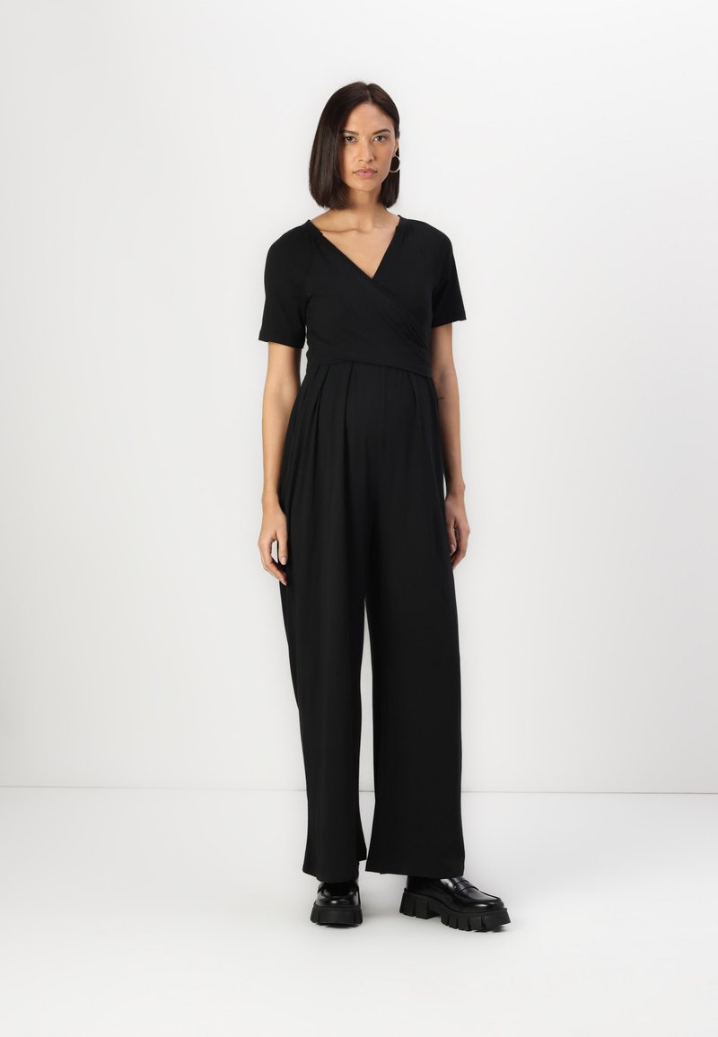 Combinaison noire avec un haut style cache-coeur et des manches courtes, présentant un pantalon large, fabriquée à partir d'un tissu lisse, associée à des chaussures noires chunkys.