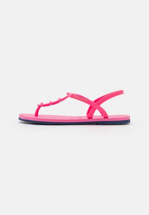 Klar pink flad sandal med tåpost, ankelrem, små dekorative nitter og tekstureret sål vist fra siden på hvid baggrund.