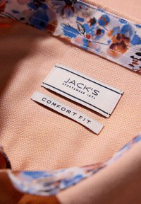 Interno di una camicia con colletto in tessuto testurizzato pesche, bordatura a motivo floreale e due etichette di stoffa: "JACK'S SPORTSWEAR INTL" e "COMFORT FIT."
