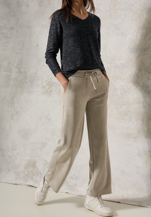 Woman wearing dark gray long-sleeve top, beige drawstring pants, and white sneakers standing against textured wall with hands in pockets.
