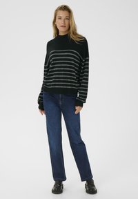 Woman standing wearing a black and gray striped sweater, blue jeans, and black loafers against a plain light background.