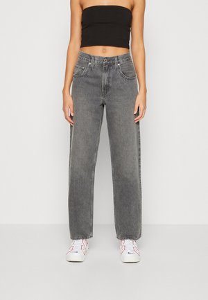 Personne portant un jean gris taille haute droit, un top tube noir et des baskets blanches, debout devant un fond clair uni.