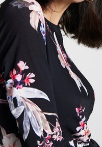 Lähikuva henkilöstä, jolla on päällä musta paita, jossa on vaaleanpunaisia, valkoisia ja purppuranvärisiä kukkakuoseja. Kuvassa näkyy olkapää sekä osittain kaula ja hiuksia.