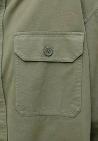 Gros plan sur une poche en tissu vert kaki avec un rabat pointu maintenu par un bouton assorti, montrant des coutures détaillées.