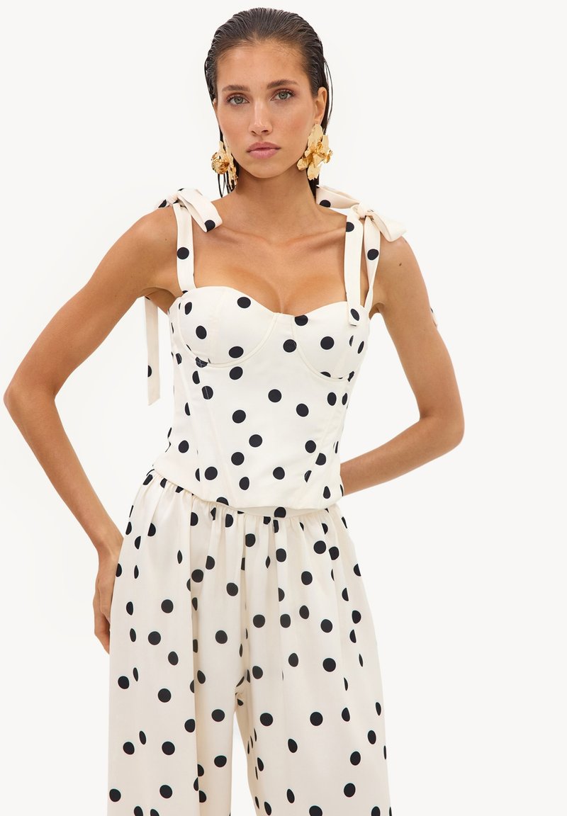 Femme en haut blanc sans manches et pantalon large à pois noirs, portant de grandes boucles d’oreilles florales dorées, debout les mains sur les hanches.