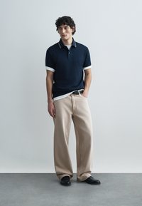 Μπλούζα πόλο ναυτικού με λευκό ριγέ γιακά, συνδυασμένη με μπεζ παντελόνι φαρδιάς γραμμής. Το μοντέλο στέκεται ουδέτερα μπροστά σε ανοιχτό φόντο.