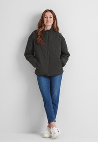 Quilted, dark green jacket with a high collar, zip front, and side pockets, paired with blue jeans and white sneakers.