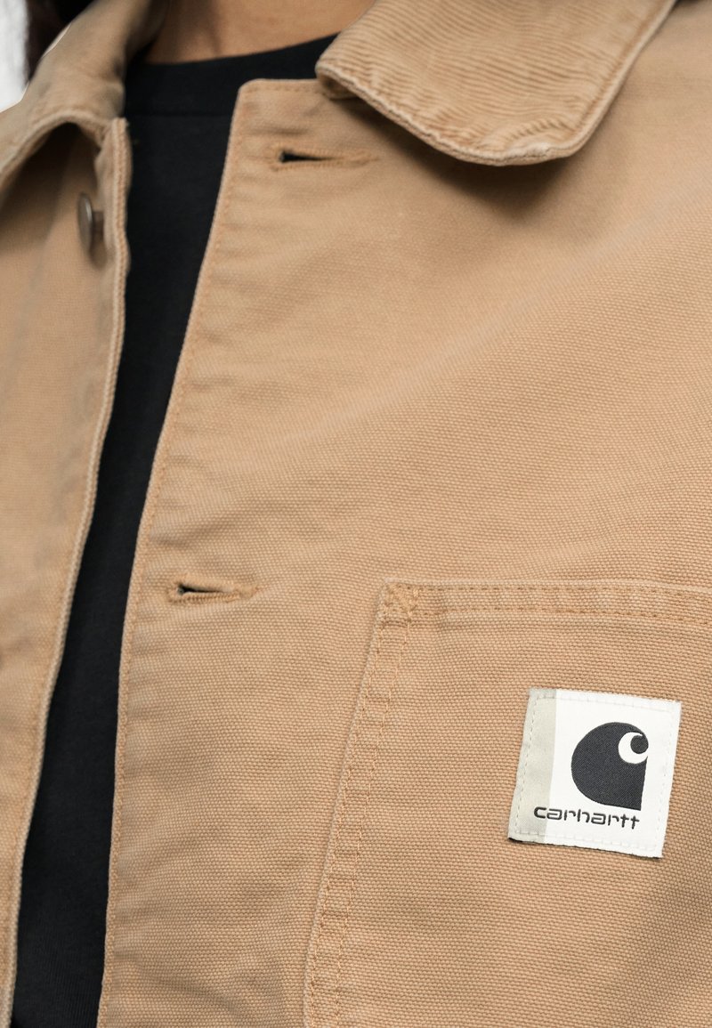 Beige canvas jas met zichtbare knoopsgaten en een vierkante Carhartt-logo patch op de borstzak, gedragen over een zwart shirt.