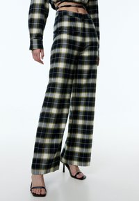 Schwarz-gelbe karierte weite Hosen mit glattem Stofftextur, hoher Taille und legerer Passform. Kombiniert mit schwarzen High Heels.