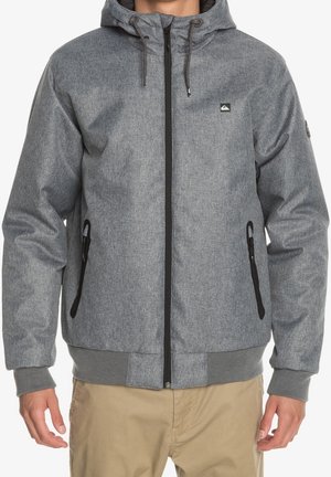 Homme portant une veste à capuche grise à fermeture éclair avec des poches zippées noires et un pantalon beige, debout devant un fond blanc.