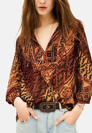 Donna che indossa una camicetta a maniche lunghe con fantasia nei toni del rosso, arancione e nero, infilata in jeans azzurri con una fibbia decorata per cintura.