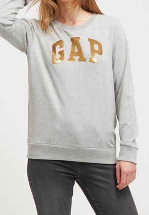 Kvinde iført lysegrå sweatshirt med store gyldne pailletbogstaver "GAP" og mørkegrå jeans, hånd hvilende på hoften.