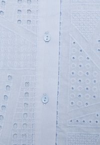Jasnoniebieska koszula z teksturowym haftem, z geometrycznymi wzorami i elementami ażurowymi. Dwa guziki widoczne wzdłuż listwy.
