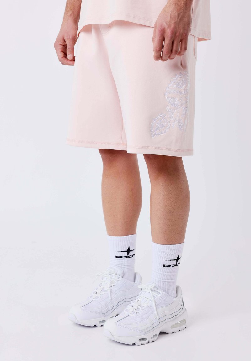 Homme portant un short rose pastel avec broderie florale, des chaussettes blanches avec logo et des baskets blanches, debout devant un fond uni.